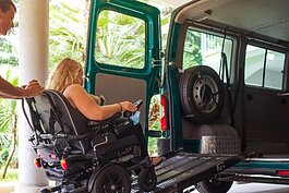 La foto muestra a una mujer en silla de ruedas siendo empujada por una rampa hacia una camioneta accesible.